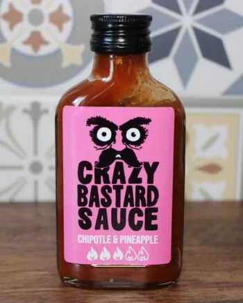 Bouteille de sauce épicée chipotle-ananas Crazy Bastard sur une table, avec des motifs colorés en arrière-plan.