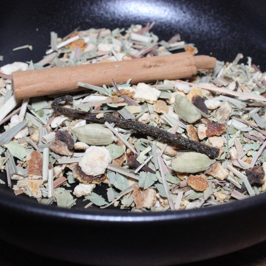 Mélange d'épices et d'herbes dans une poêle noire, comprenant bâton de cannelle, cardamome et écorces d'orange.