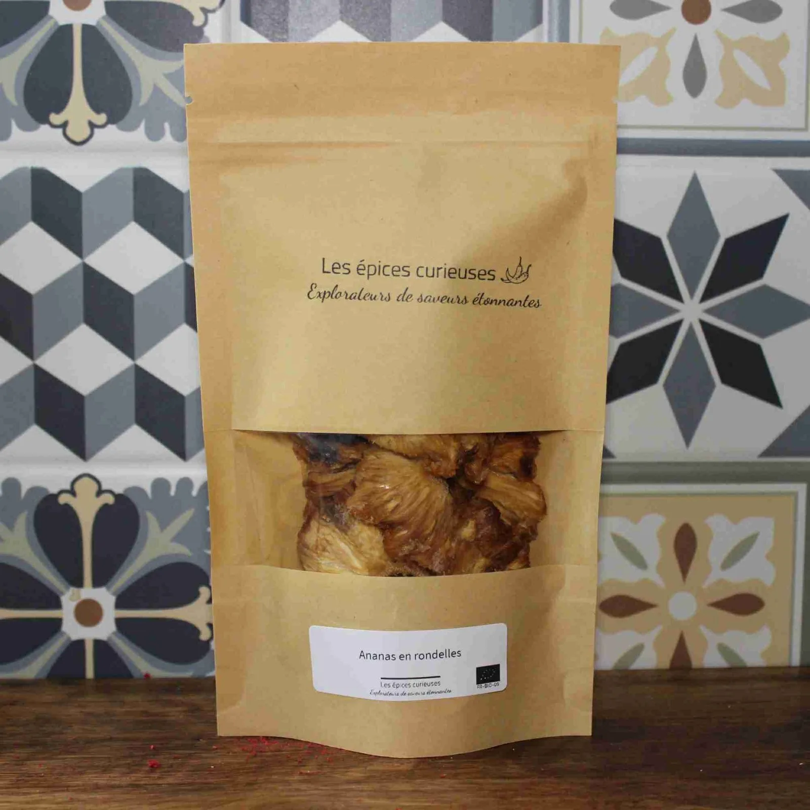 Sachet de rondelles d'ananas séchées, produit par Les Épices Curieuses, devant un carrelage coloré.