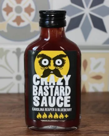 Bouteille de sauce épicée Crazy Bastard goût Carolina Reaper et myrtille sur fond de carreaux colorés.