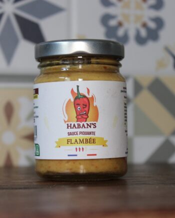 Bocal de sauce piquante Haban's flambée posé sur une table en bois avec motif de fond géométrique.