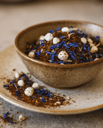 Bol de thé en vrac aux fleurs bleues et ingrédients naturels, sur assiette en céramique.